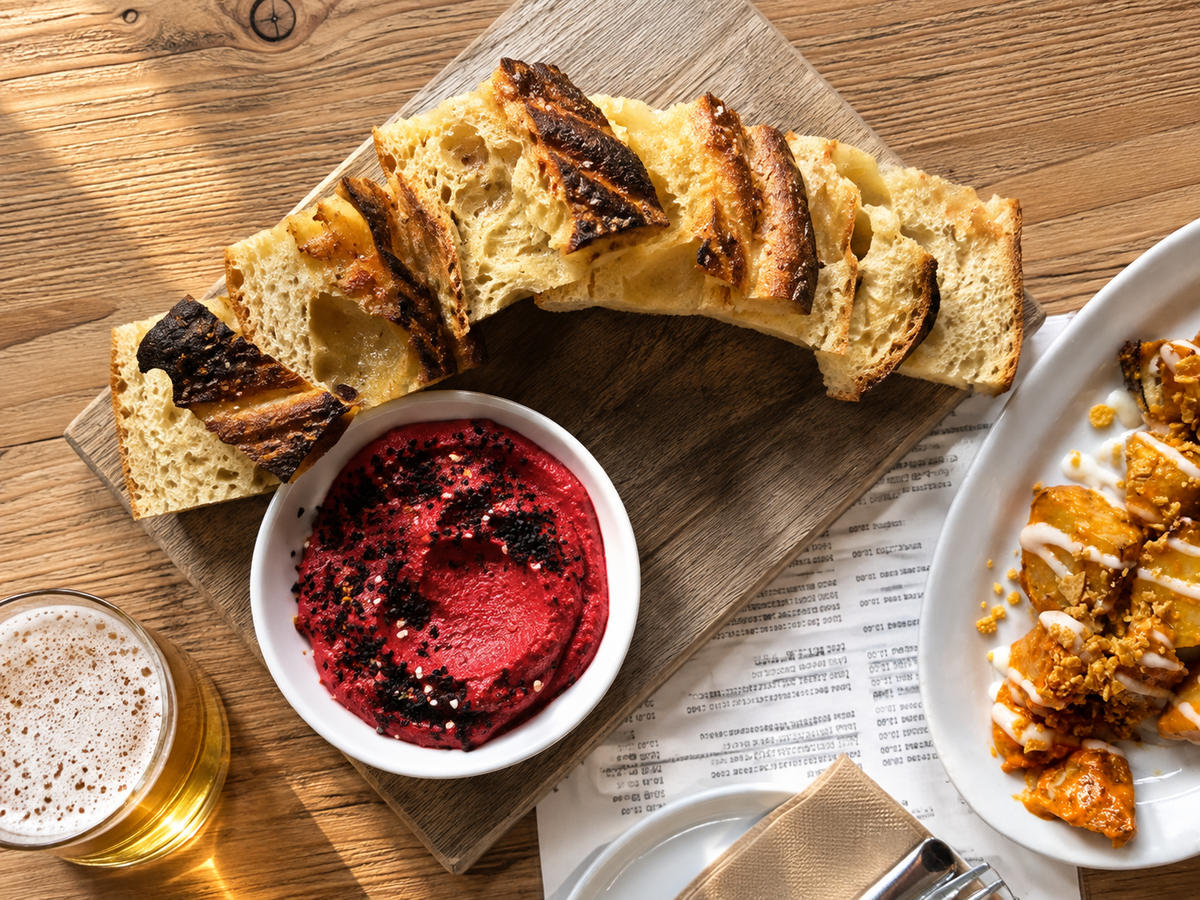A styled food scene on a wooden table showing a rustic baguette sliced and arranged on a wooden board, a small white bowl of bright red beet hummus, a glass of beer, and part of another plated dish. Delicious food at a local Victoria restaurant