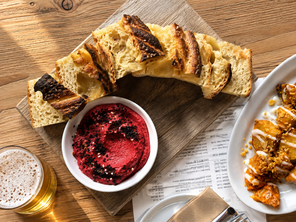 A styled food scene on a wooden table showing a rustic baguette sliced and arranged on a wooden board, a small white bowl of bright red beet hummus, a glass of beer, and part of another plated dish. Delicious food at a local Victoria restaurant