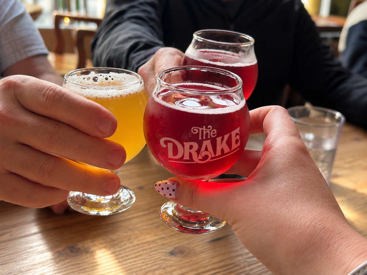 Three people clinking taster glasses in a toast at a wooden table. The center glass is branded with 'the DRAKE' in white script and contains a red drink, while the other two glasses contain yellow and pinkish-red beverages.
