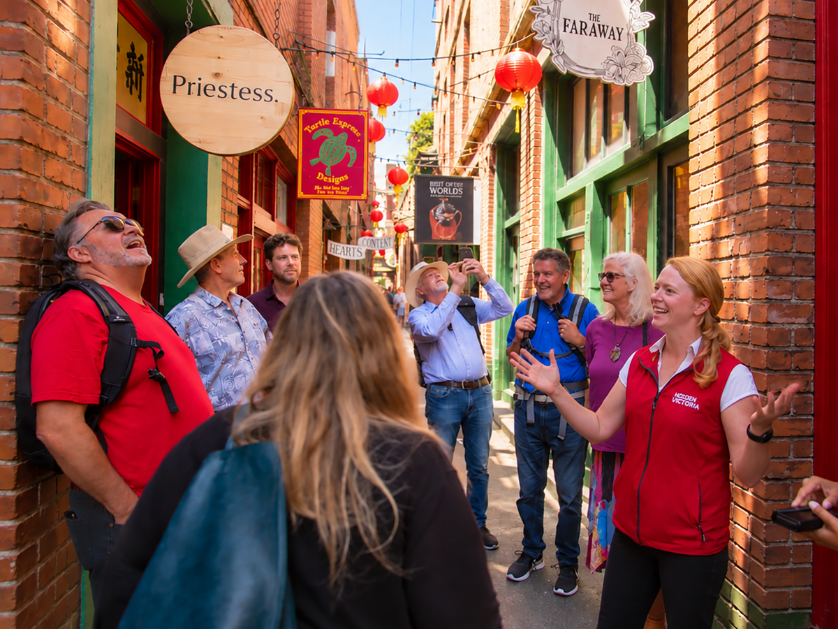 A group of tourists listening to a guide who wears a red and white branded vest while standing in Fan Tan Alley, lively street lined with brick buildings and colourful shop signs.