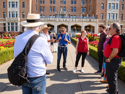 A group of tourists stands outdoors in front of The Empress hotel, listening to a guide who is holding a camera. The scene is set on a sunny day with a clear view of the historic building and flower beds in the background.