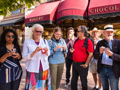 A group of seven people standing on a sidewalk in front of Rogers' chocolate shop with red awnings. Several people are holding and eating small pink and red sweets or chocolates.